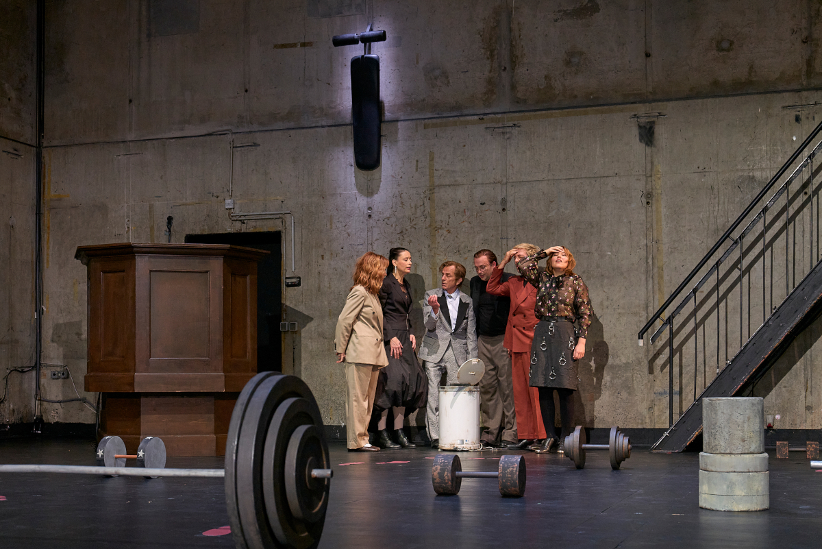 Das Ensemble steht in einem Halbkreis um einen ge&ouml;ffneten wei&szlig;en Eimer herum und interagiert miteinander. Josefine Israel und Samuel Weiss fassen sich mit einer Hand an den Kopf. Im B&uuml;hnenbild liegen verschiedene rostige Kurz- und Langhanteln, links befindet sich eine h&ouml;lzerne Kanzel. An der Betonwand im Hintergrund h&auml;ngt eine Hantelbank, die mit einem Spot beleuchtet ist.